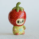 Small ceramic sculpture of a character wearing a giant strawberry around their head. They sit with a tiny body.