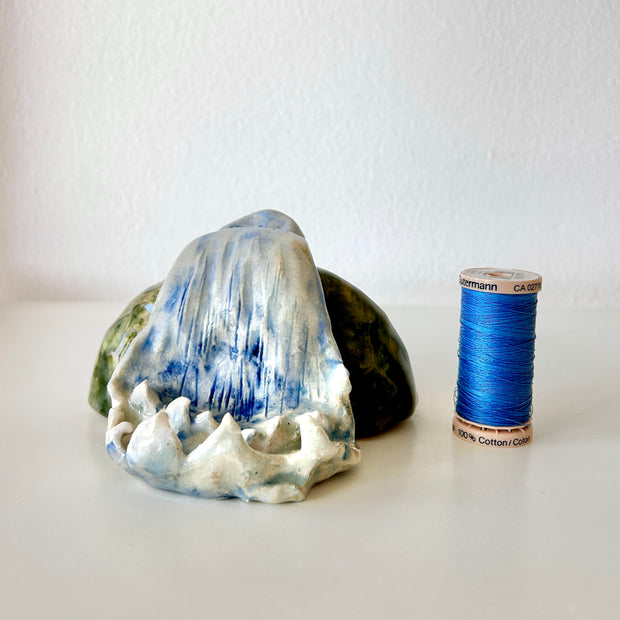 Ceramic sculpture of a waterfall over a rock. Sculpture is next to a spool of thread for size reference.