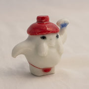 Porcelain sculpture of the radish spirit from Spirited Away, standing in a red string bottom and with a red hat. It holds up a fan in one hand.