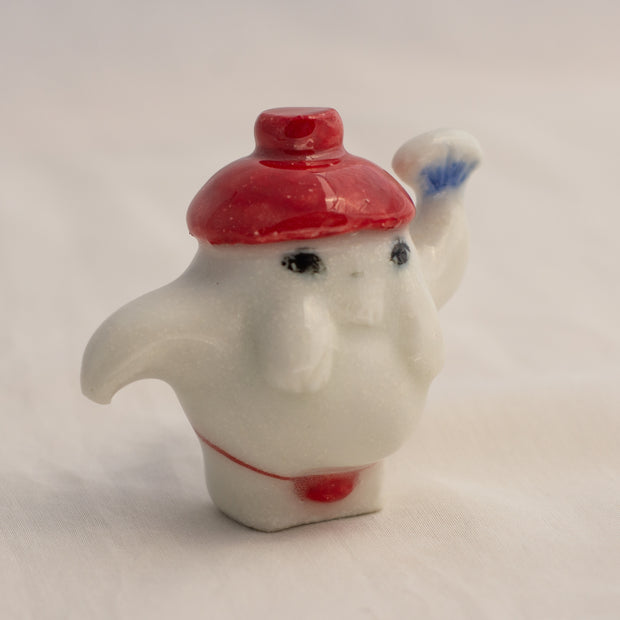 Porcelain sculpture of the radish spirit from Spirited Away, standing in a red string bottom and with a red hat. It holds up a fan in one hand.