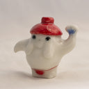 Porcelain sculpture of the radish spirit from Spirited Away, standing in a red string bottom and with a red hat. It holds up a fan in one hand.