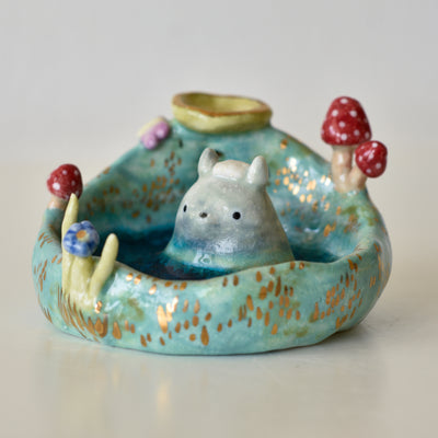 Ceramic sculpture of Totoro half submerged in an outdoor hot spring onsen, with a towel on its head and surrounded by mushrooms.