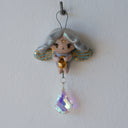 Hanging ceramic figure of a cartoon, cute fairy with wings and gold detailings. Hanging from her is a colorful crystal.