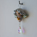 Hanging ceramic figure of a cartoon, cute fairy with wings and gold detailings. Hanging from her is a colorful crystal.