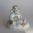 Small figurine of a fairy on a rainbow with stars and a very small pink bunny, all positioned on a fluffy cloud-like base. 