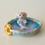 Ceramic sculpture of a creature that is half mermaid, half bunny, resting in a rounded body of water with a small gold crescent moon attached.