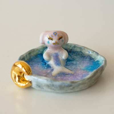 Ceramic sculpture of a creature that is half mermaid, half bunny, resting in a rounded body of water with a small gold crescent moon attached.
