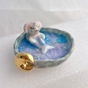 Ceramic sculpture of a creature that is half mermaid, half bunny, resting in a rounded body of water with a small gold crescent moon attached.
