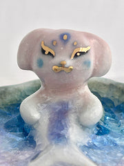 Ceramic sculpture of a creature that is half mermaid, half bunny, resting in a rounded body of water with a small gold crescent moon attached.