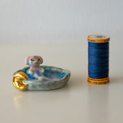 Ceramic sculpture of a creature that is half mermaid, half bunny, resting in a rounded body of water with a small gold crescent moon attached.