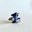 Tiny ceramic sculpture of a bird with a gold beak and gold and black wings, wearing a blue cloak and holding a small cane.