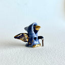 Tiny ceramic sculpture of a bird with a gold beak and gold and black wings, wearing a blue cloak and holding a small cane.