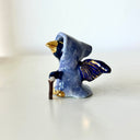 Tiny ceramic sculpture of a bird with a gold beak and gold and black wings, wearing a blue cloak and holding a small cane.