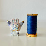 Small ceramic rabbit figure next to a spool of blue thread on a light background