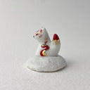 Small ceramic figurine of a white fox with red and gold color accents, sitting atop of a cloud like shape.