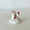 Small ceramic figurine of a white fox with red and gold color accents, sitting atop of a cloud like shape.