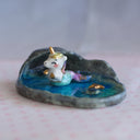 Ceramic sculpture of a water cover, framed with rocks. A colorful unicorn mermaid sits atop the water (top half is a unicorn, bottom is a mermaid tail).