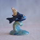 Ceramic sculpture of a small white bunny riding atop of a blue fish like a mount, which is positioned atop of a tall wave.