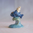 Ceramic sculpture of a small white bunny riding atop of a blue fish like a mount, which is positioned atop of a tall wave.