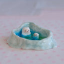 Ceramic sculpture of a pool surrounded by a wall of ice and snow, inside the pool sit 2 small white yeti characters with smiling faces.