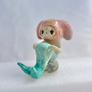 Ceramic figurine of a animal wearing a pink bunny hood hat, they have a mermaid tail and sit on a rock.