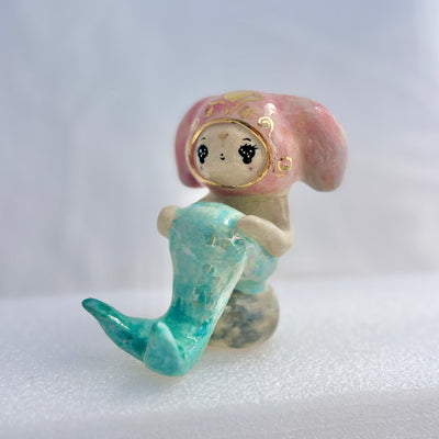 Ceramic figurine of a animal wearing a pink bunny hood hat, they have a mermaid tail and sit on a rock.