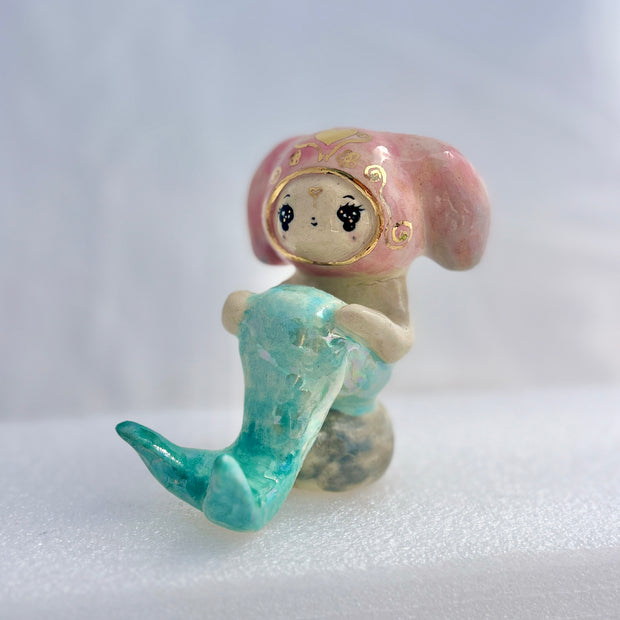 Ceramic figurine of a animal wearing a pink bunny hood hat, they have a mermaid tail and sit on a rock.