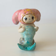 Ceramic figurine of a animal wearing a pink bunny hood hat, they have a mermaid tail and sit on a rock.