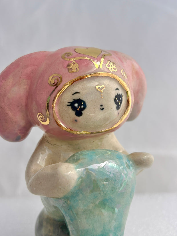 Ceramic figurine of a animal wearing a pink bunny hood hat, they have a mermaid tail and sit on a rock.
