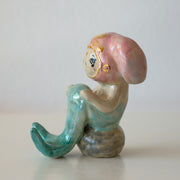 Ceramic figurine of a animal wearing a pink bunny hood hat, they have a mermaid tail and sit on a rock.
