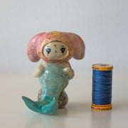 Ceramic figurine of a animal wearing a pink bunny hood hat, they have a mermaid tail and sit on a rock.