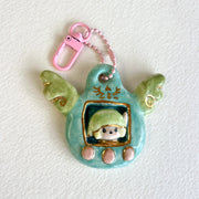 Ceramic sculpture attached to a keychain, sculpture features a small figurine inside a green tamagotchi-like frame with pink and gold color accents on a white background