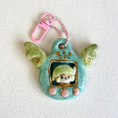 Ceramic sculpture attached to a keychain, sculpture features a small figurine inside a green tamagotchi-like frame with pink and gold color accents on a white background