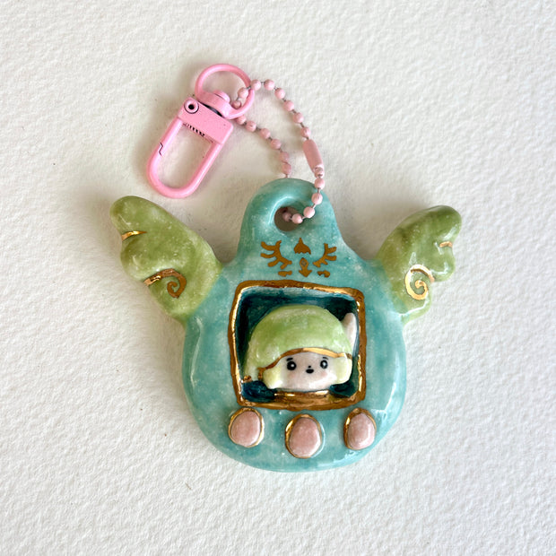 Ceramic sculpture attached to a keychain, sculpture features a small figurine inside a green tamagotchi-like frame with pink and gold color accents on a white background