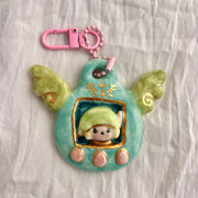 Ceramic sculpture attached to a keychain, sculpture features a small figurine inside a green tamagotchi-like frame with pink and gold color accents on a white background