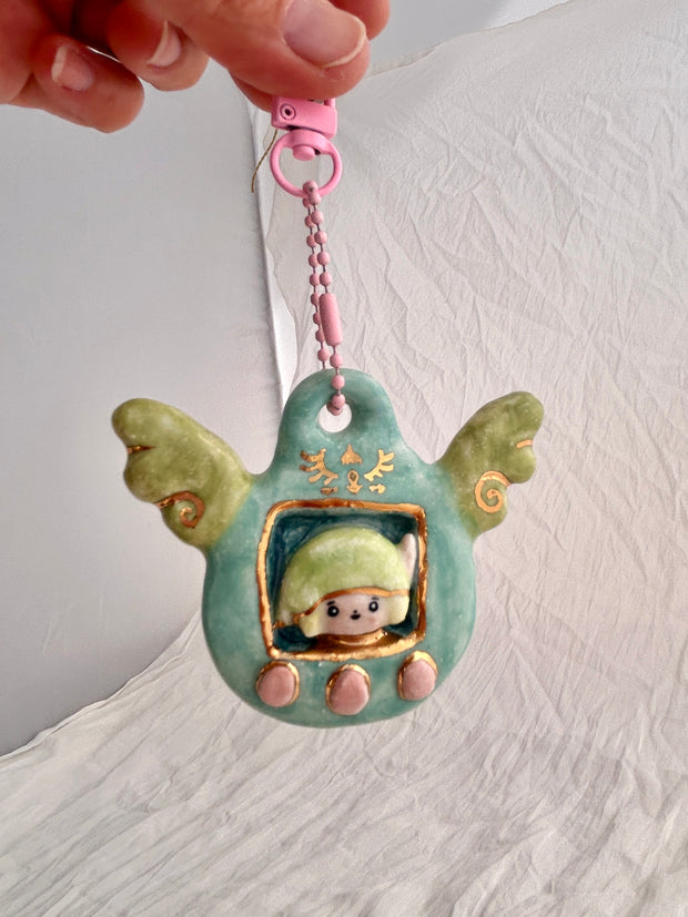 Ceramic sculpture attached to a keychain, sculpture features a small figurine inside a green tamagotchi-like frame with pink and gold color accents on a white background