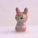 Pink rabbit figurine with a yellow heart on its forehead. It sits on a light pink background