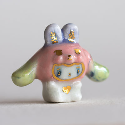 Ceramic white bunny figurine with a pink bunny hood hat with gold and green accents on a light gray background