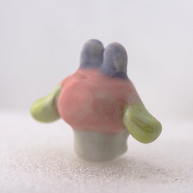 Ceramic white bunny figurine with a pink bunny hood hat with gold and green accents on a light gray background