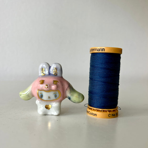 Ceramic white bunny figurine with a pink bunny hood hat with gold and green accents on a light gray background, it sits next to a spool for size reference.