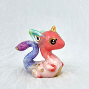 Colorful dragon-like figurine with pink body, purple tail, and gold accents on a light gray background