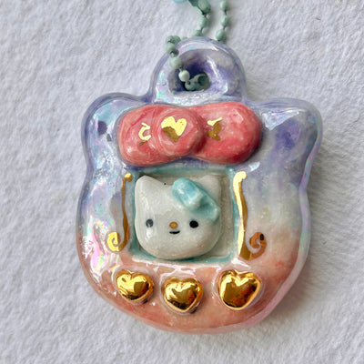 Ceramic sculpture of a Tamagotchi with gold buttons and a Hello Kitty like character in the screen.