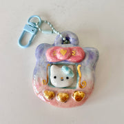 Ceramic keychain sculpture of a Tamagotchi with gold buttons and a Hello Kitty like character in the screen.