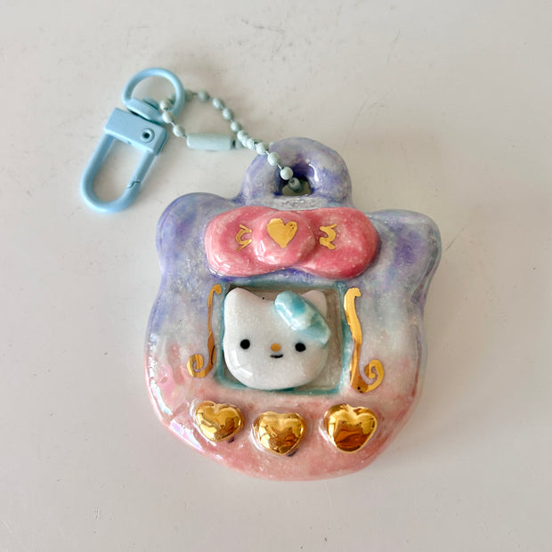 Ceramic keychain sculpture of a Tamagotchi with gold buttons and a Hello Kitty like character in the screen.