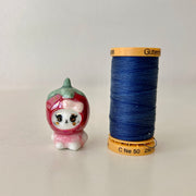 Small figurine of a bear in a strawberry costume next to a spool of blue thread on a light gray background