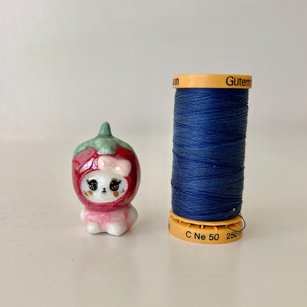 Small figurine of a bear in a strawberry costume next to a spool of blue thread on a light gray background