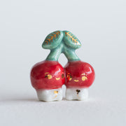 Ceramic sculpture of 2 tiny, cute cherry characters connected to the same stem. They have gold detailing.