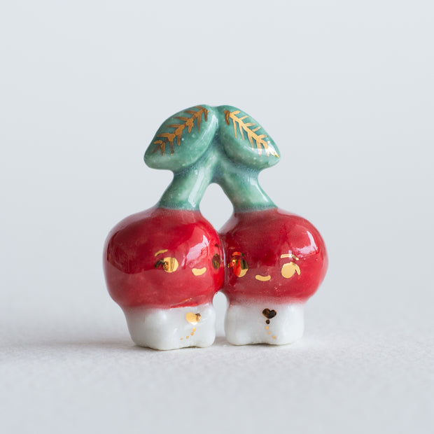 Ceramic sculpture of 2 tiny, cute cherry characters connected to the same stem. They have gold detailing.