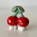 Ceramic sculpture of 2 tiny, cute cherry characters connected to the same stem. They have gold detailing.