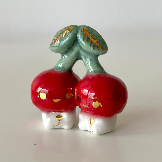 Ceramic sculpture of 2 tiny, cute cherry characters connected to the same stem. They have gold detailing.